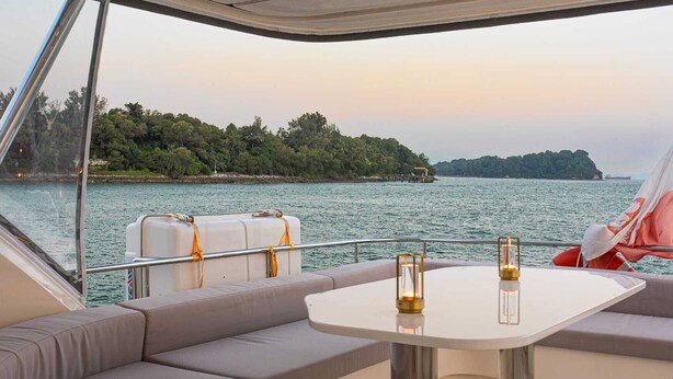 Interior de barco com área de estar acolchoada, uma pequena mesa com duas velas e vista para uma ilha arborizada através da água.