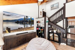 A living room with a large flat-screen TV displaying a scenic lake view, wooden flooring, a cozy bean bag, a dark staircase, and a kitchen area in the background. - Glenn Getaway - Nicely Remodeled (Tahoe Vista)