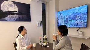 Two people seated at a round table with a cityscape TV on the wall. - Moon Base Tokyo Inn (Tokyo)
