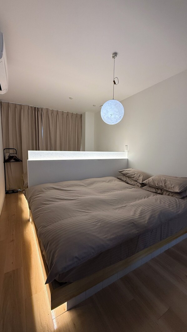 Ett sovrum med en säng, en hängande lampa, ett fönster med gardiner och ett trägolv.