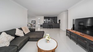 A modern living room with a sofa, a coffee table with flowers, a TV, and a kitchen in the background. - Blanco Haus - Sweet Family Escape near Theme Parks (Pimpama)