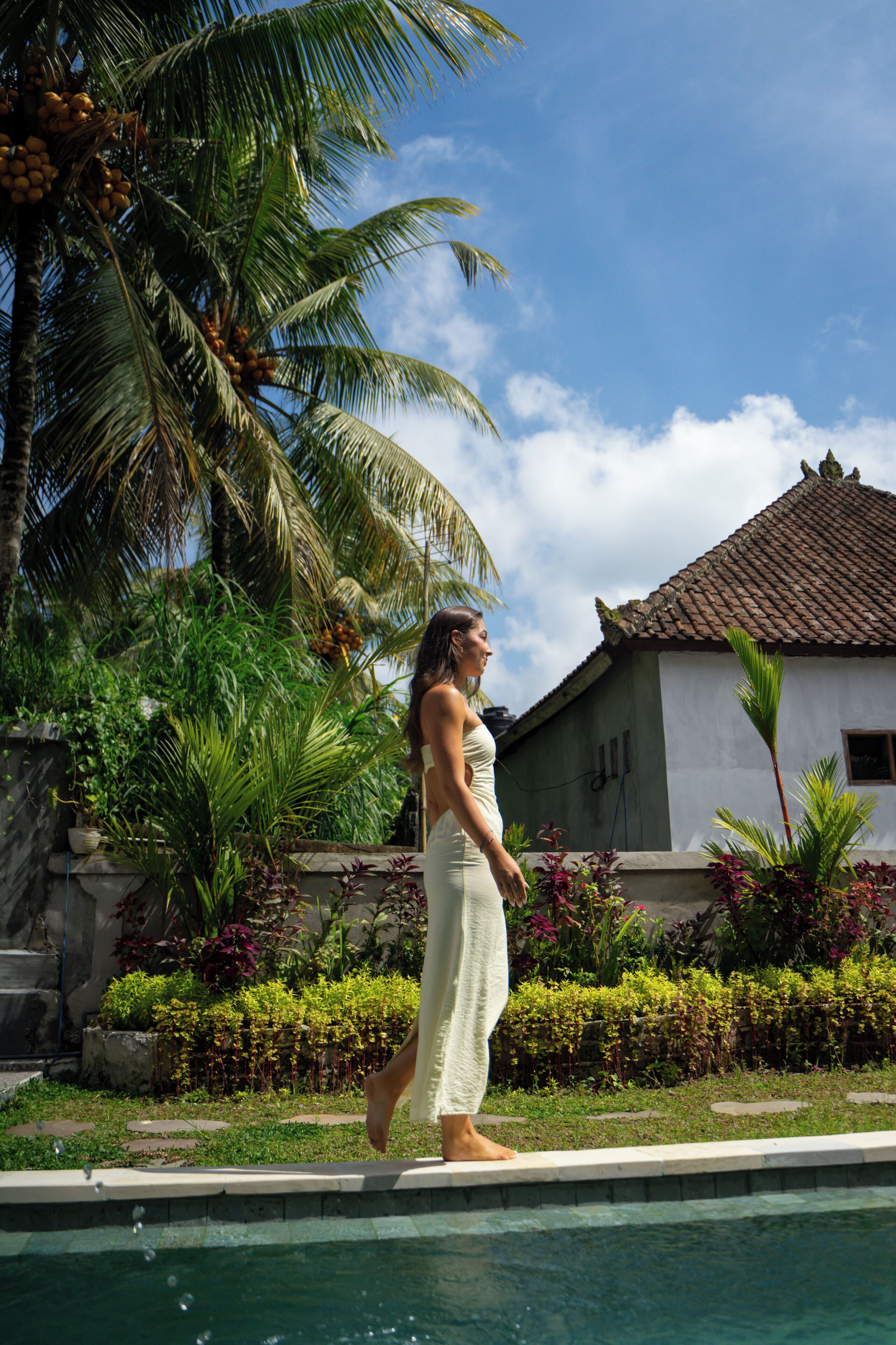 A woman standing by a pool with palm trees and a house in the background.