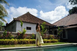 Mulher de vestido branco parada perto de uma piscina, com uma casa ao fundo.