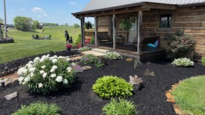 A log cabin with a porch, surrounded by landscaped garden beds with various plants and flowers.