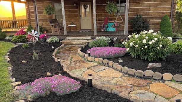 A log cabin with a stone pathway leading to the entrance, surrounded by flower beds and a wooden deck.