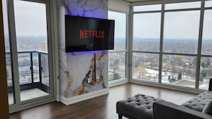 A spacious room with a large window offering a city view, a flat-screen TV mounted on a marble wall, and a comfortable seating area with a cushioned bench. - Luxury Condo Toronto YYZ (Toronto)