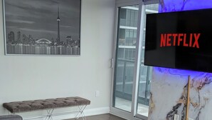 A modern living room with a grey sectional sofa, a bench, and a ceiling fan. A large TV screen displays the Netflix logo. - Luxury Condo Toronto YYZ (Toronto)