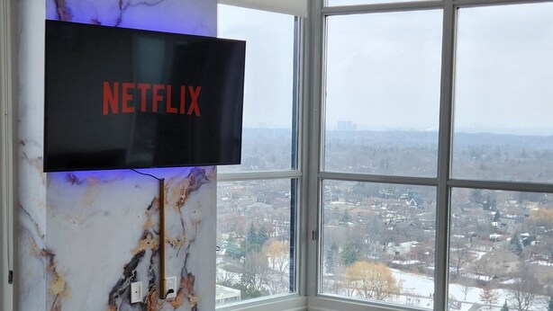 A modern living room with a large TV mounted on a marble wall, a dark wood floor, and a view of a cityscape through large windows. - Luxury Condo Toronto YYZ (Toronto)