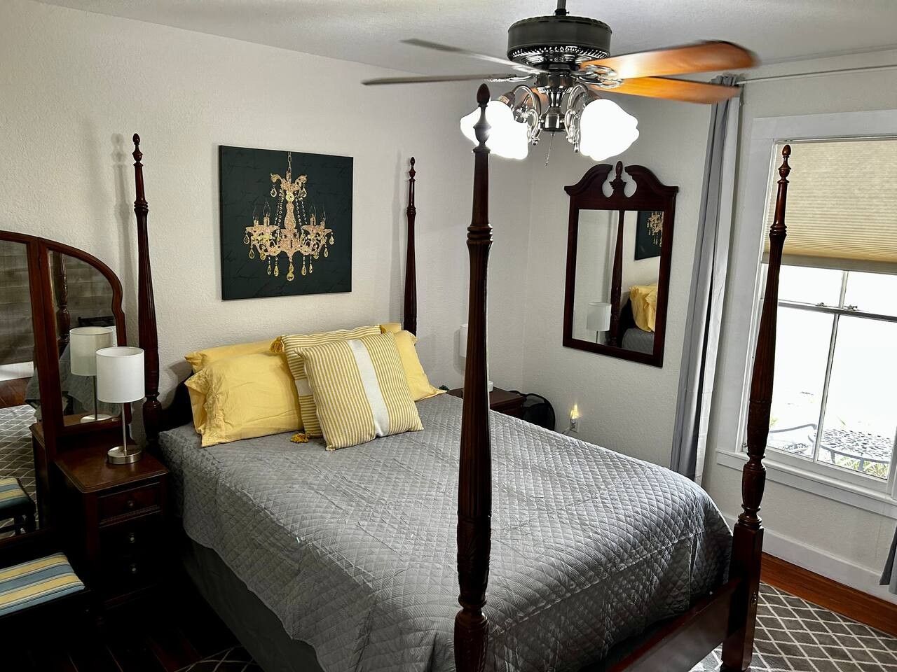 A four-poster bed with a chandelier, a bedside table with a lamp, a mirror, and a window with blinds.