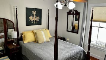 A four-poster bed with a chandelier, a bedside table with a lamp, a mirror, and a window with blinds.