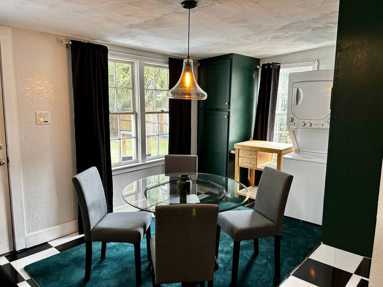 A room with a glass table surrounded by chairs, a hanging light fixture, a window with curtains, and a kitchen area with a washing machine.