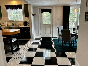 Una habitación con piso a cuadros, una mesa de comedor con sillas, una zona de cocina con pileta y armarios, y una puerta con vista al exterior.