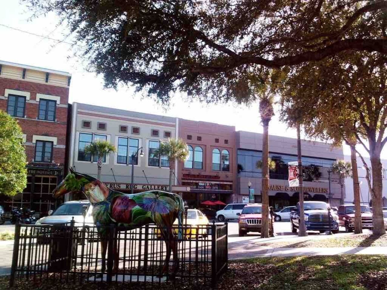 A street scene with a colorful horse statue in the foreground, parked cars, and buildings with storefronts in the background.