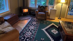 A cozy living room with a wood-burning stove, a leather armchair, a patterned rug, and a glass display case. - Charming Log Home on acreage bordering golf course with full gym (Port Alberni)