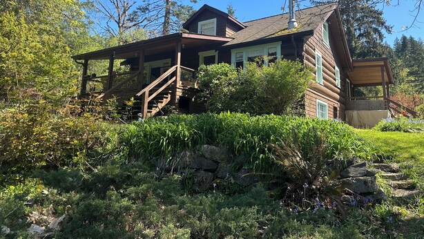A log cabin with a porch, a chimney, and a stone wall. - Charming Log Home on acreage bordering golf course with full gym (Port Alberni)