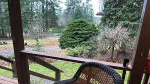 A wicker chair on a wooden deck with a view of a forested area and a road. - Charming Log Home on acreage bordering golf course with full gym (Port Alberni)