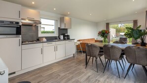 Een moderne keuken met witte kastjes, een ingebouwde oven en een koffiezetapparaat. Er is een eethoek met een tafel en stoelen, en een groot raam met uitzicht op de omgeving.