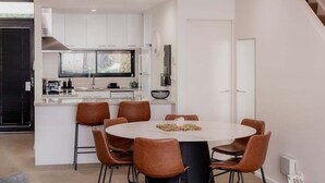 A modern kitchen with white cabinets, a central island, and a dining area with brown chairs. - Heart of Hobart | CBD Apartment & Parking (Hobart City)