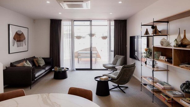A modern living room with a sofa, armchair, coffee table, and a dining area. There is a bookshelf with decorative items and a large window with curtains. - Heart of Hobart | CBD Apartment & Parking (Hobart City)