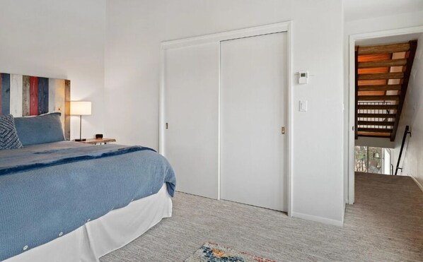 A bedroom with a bed, a nightstand, a lamp, a door, and a view of a wooden staircase.