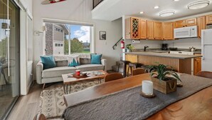 A modern kitchen with wooden cabinets, a microwave, and a sink. There is a dining area with a table and chairs, and a sofa with cushions.