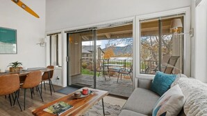 A modern living room with a sofa, coffee table, and dining area. There is a sliding glass door leading to a balcony with outdoor furniture.