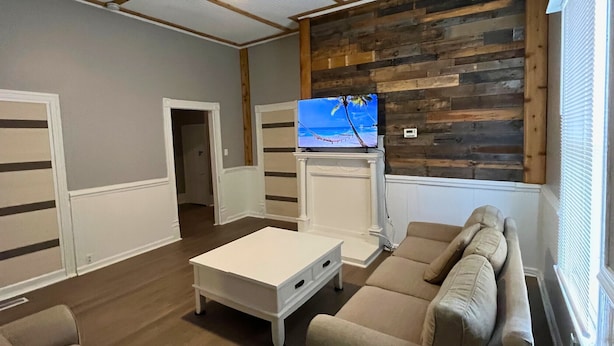 Uma sala de estar com uma mesa de centro branca, sofá bege e uma televisão em uma parede de madeira.