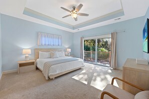 Ein Schlafzimmer mit Bett, Nachttischen, Deckenventilator, großem Fenster mit Blick ins Grüne und einer Schiebetür.