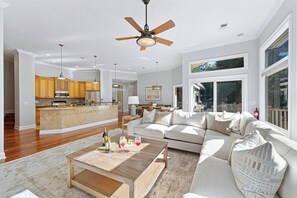 A spacious living area with a large sofa, a coffee table, and a ceiling fan. There is a kitchen area with wooden cabinets and a dining table. - Pet Friendly Palmetto Dunes Home w/ Private Pool, Game Room & Golf Views (Hilton Head Island)