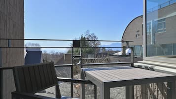 Un balcón con mesa y silla de madera, baranda de vidrio y vistas a edificios y montañas.