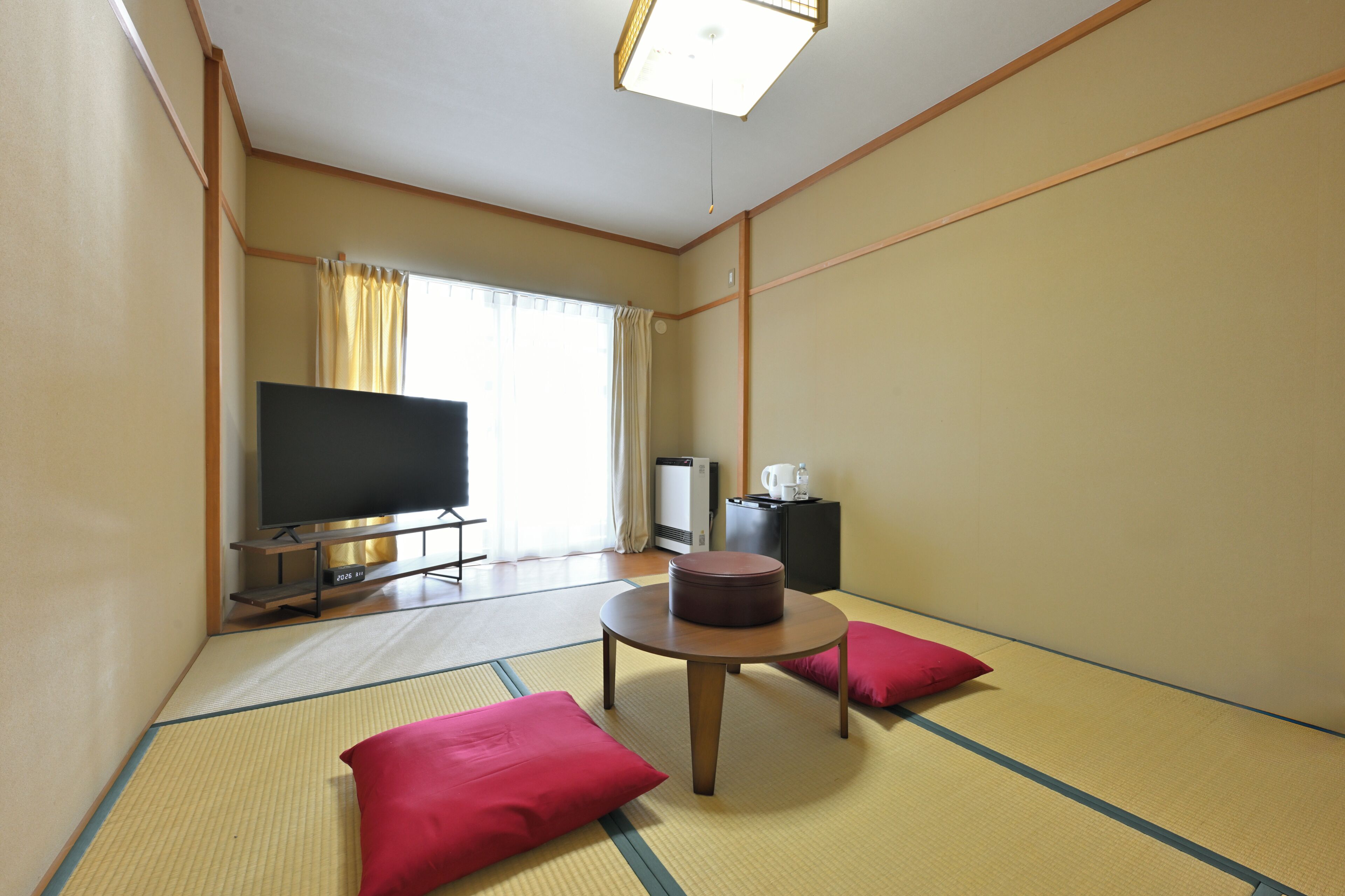 Ein Zimmer mit Tatami-Boden, einem Fernseher, einem kleinen Tisch und einer Couch mit Kissen.