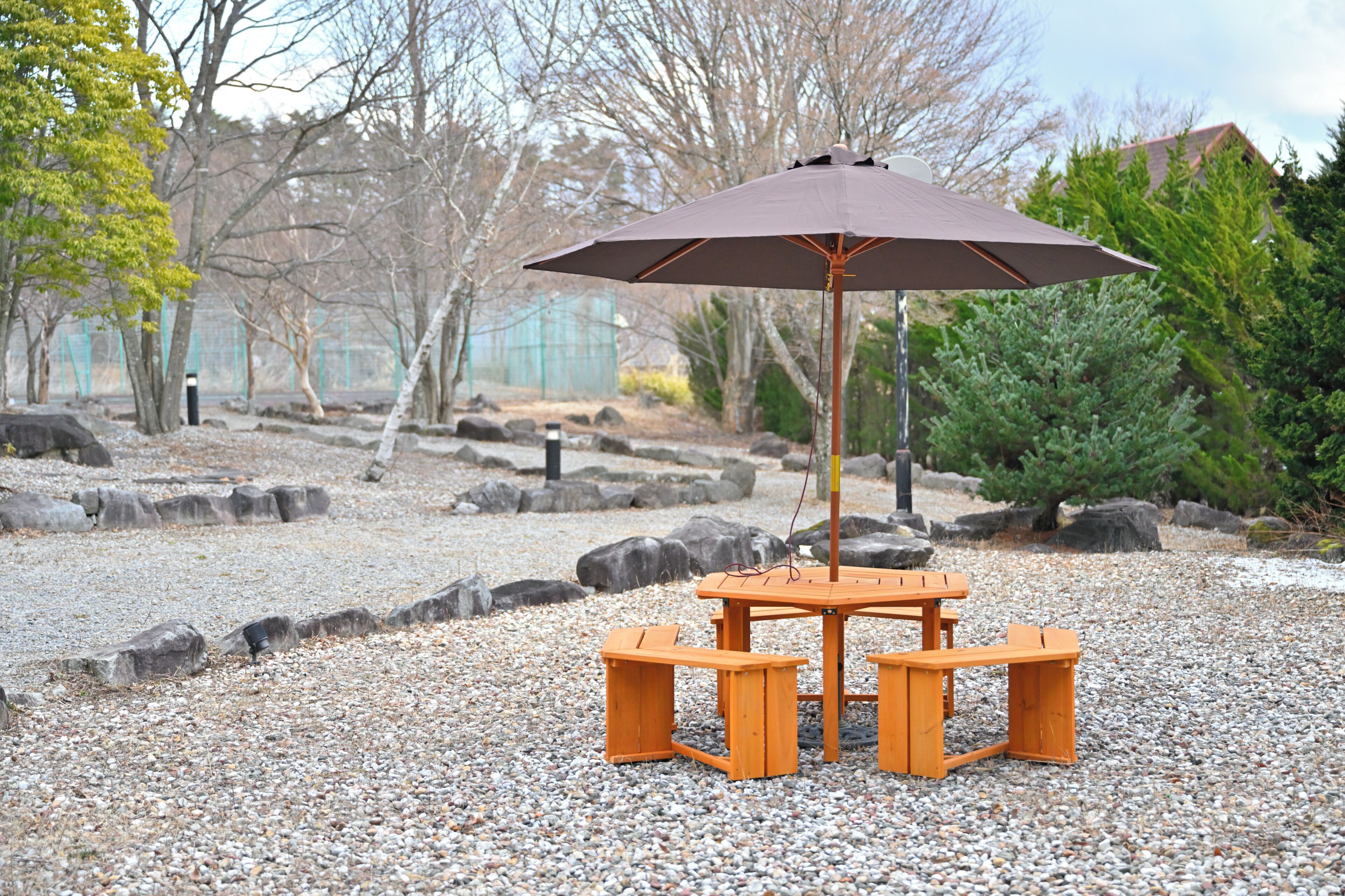 A wooden table and bench set with an umbrella in a gravel area surrounded by rocks and trees.