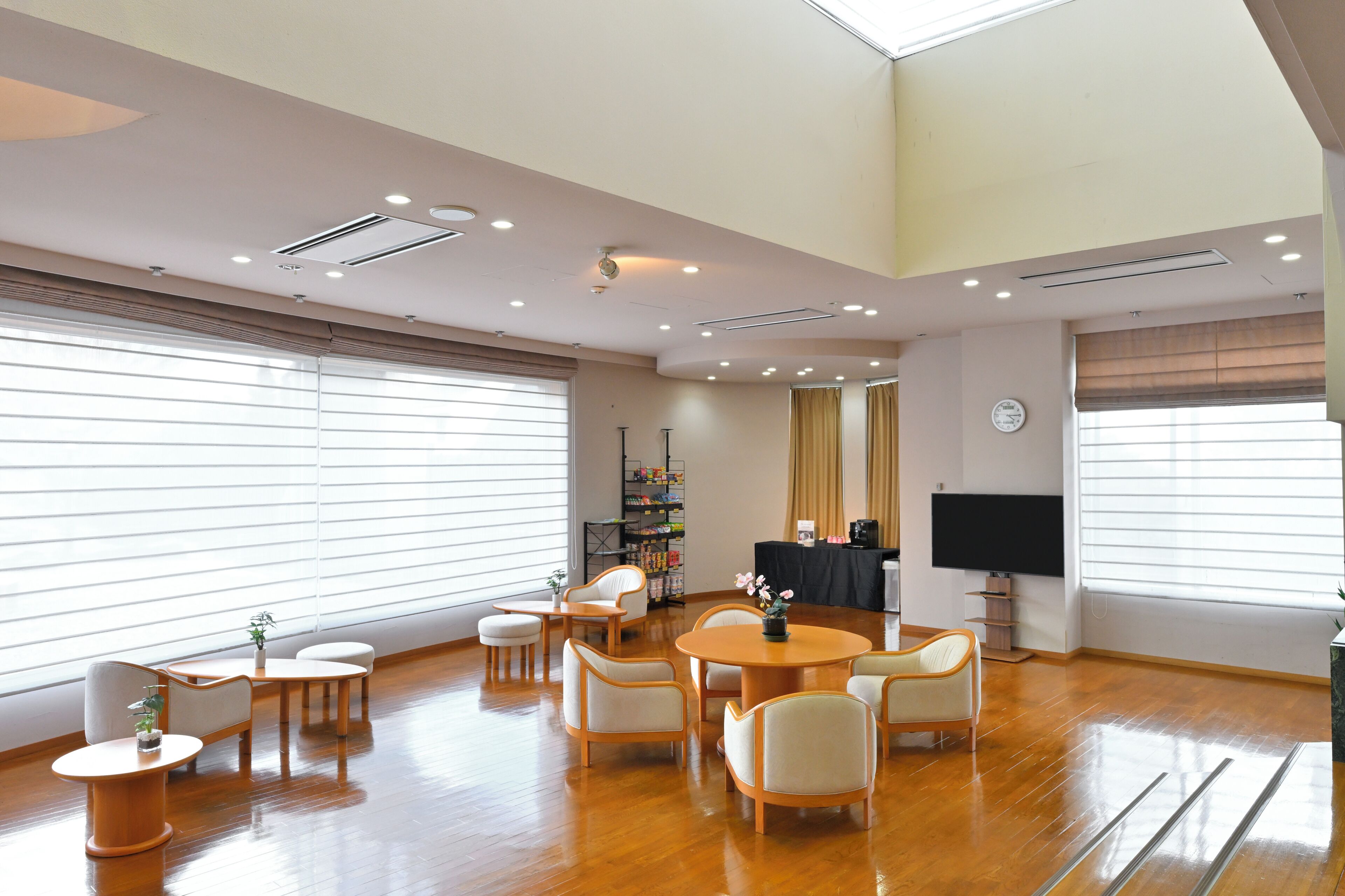A spacious room with a high ceiling, wooden flooring, a round table with chairs, a bookshelf, a television, and large windows with blinds.