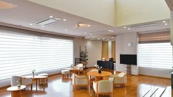 A spacious room with a high ceiling, wooden flooring, a round table with chairs, a bookshelf, a television, and large windows with blinds.