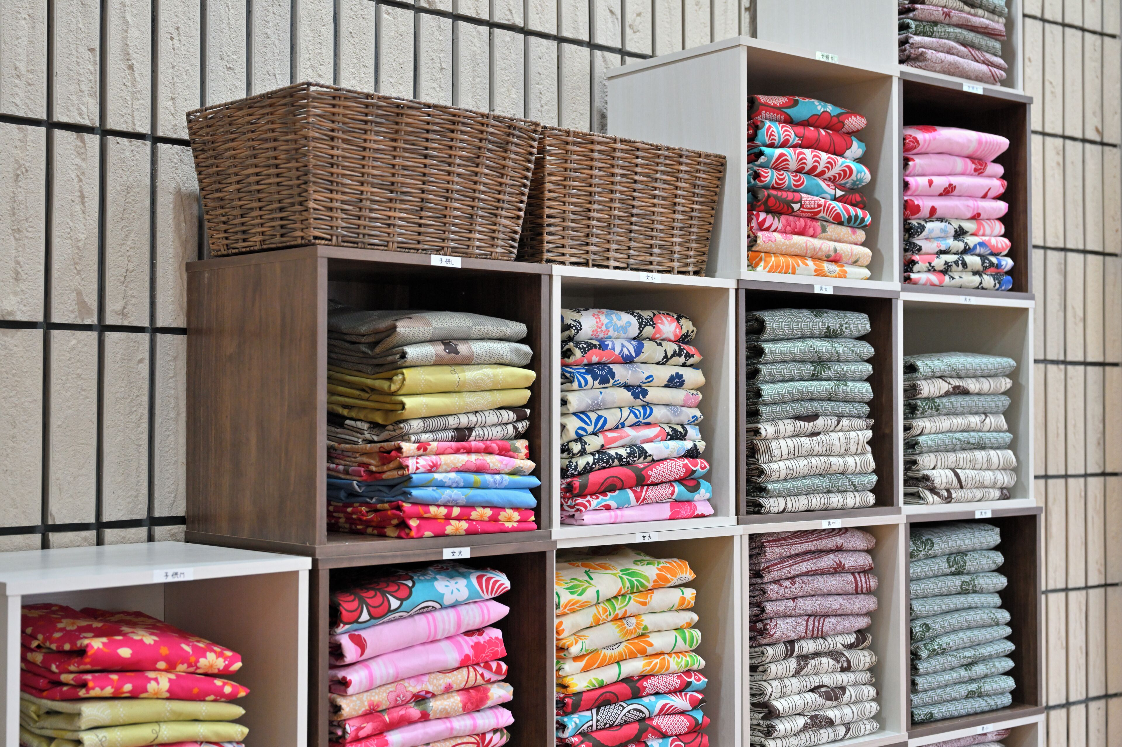 A display of neatly folded fabrics in various colors and patterns, organized on shelves against a textured wall.