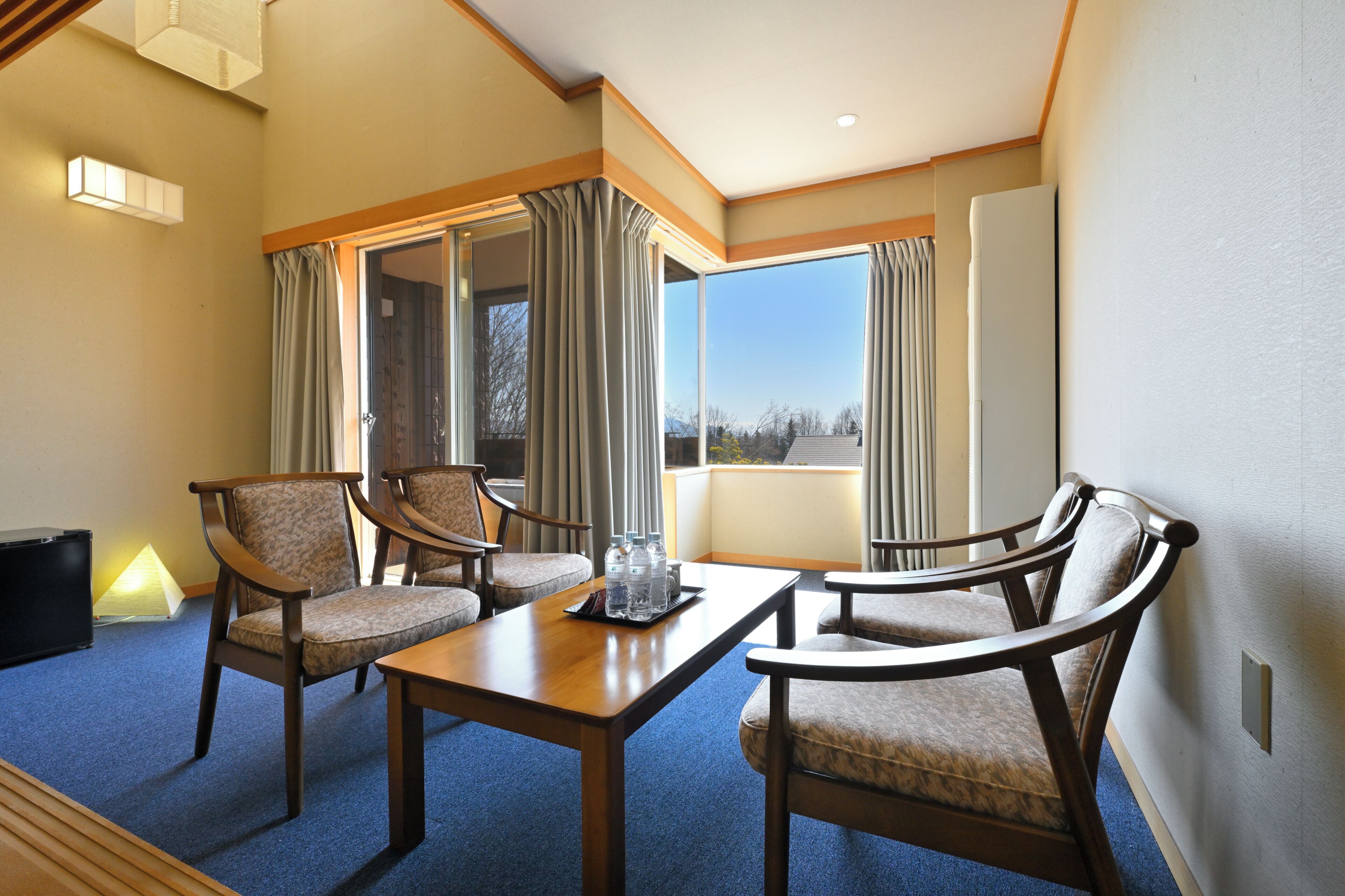 A hotel room with a large window, a dining table with chairs, and a lamp on the floor.