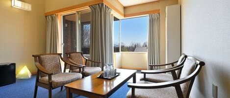 A hotel room with a large window, a dining table with chairs, and a lamp on the floor.