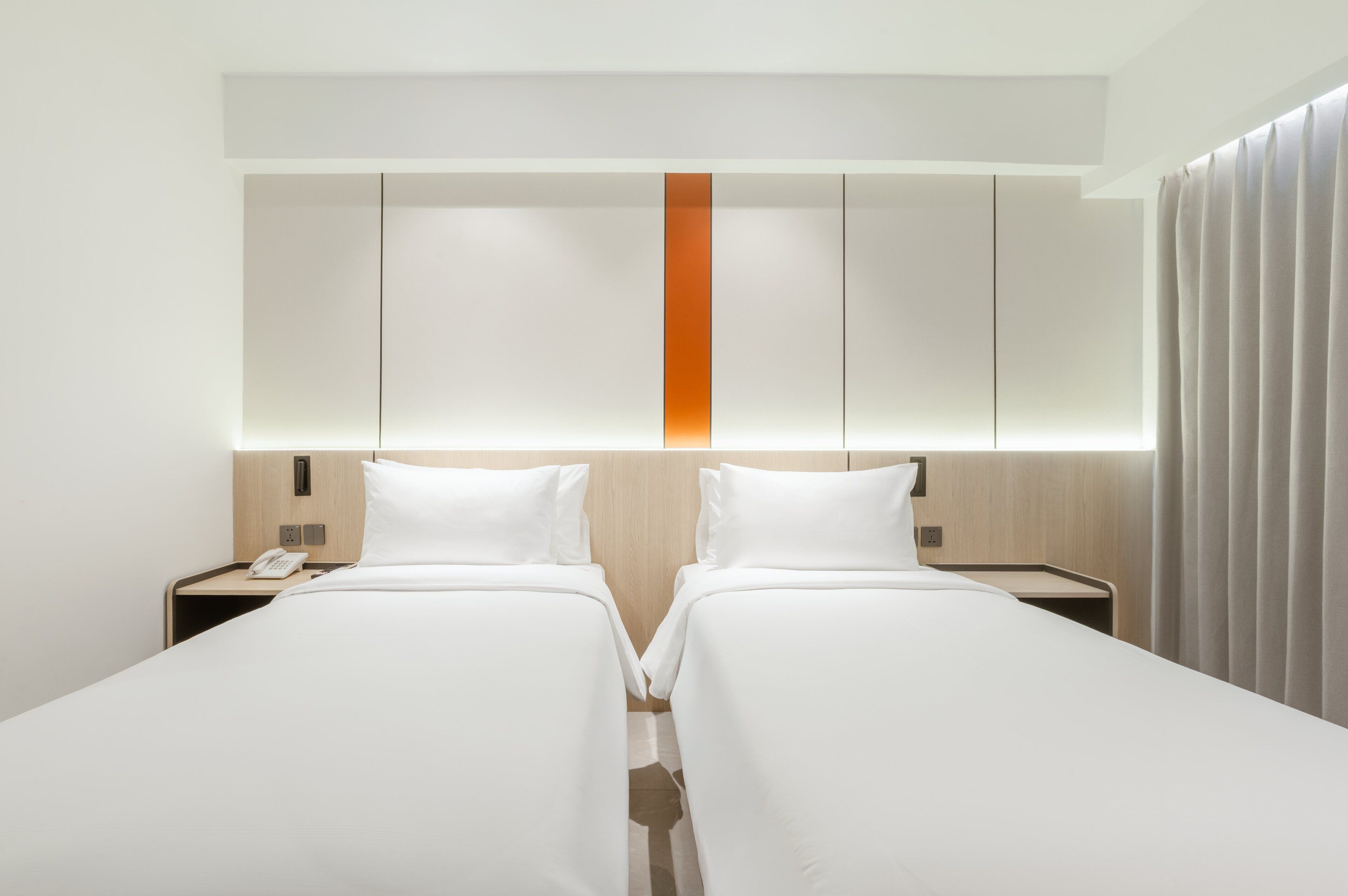 A hotel room with two single beds, a wooden headboard with a vertical orange accent, and a bedside table with a phone.