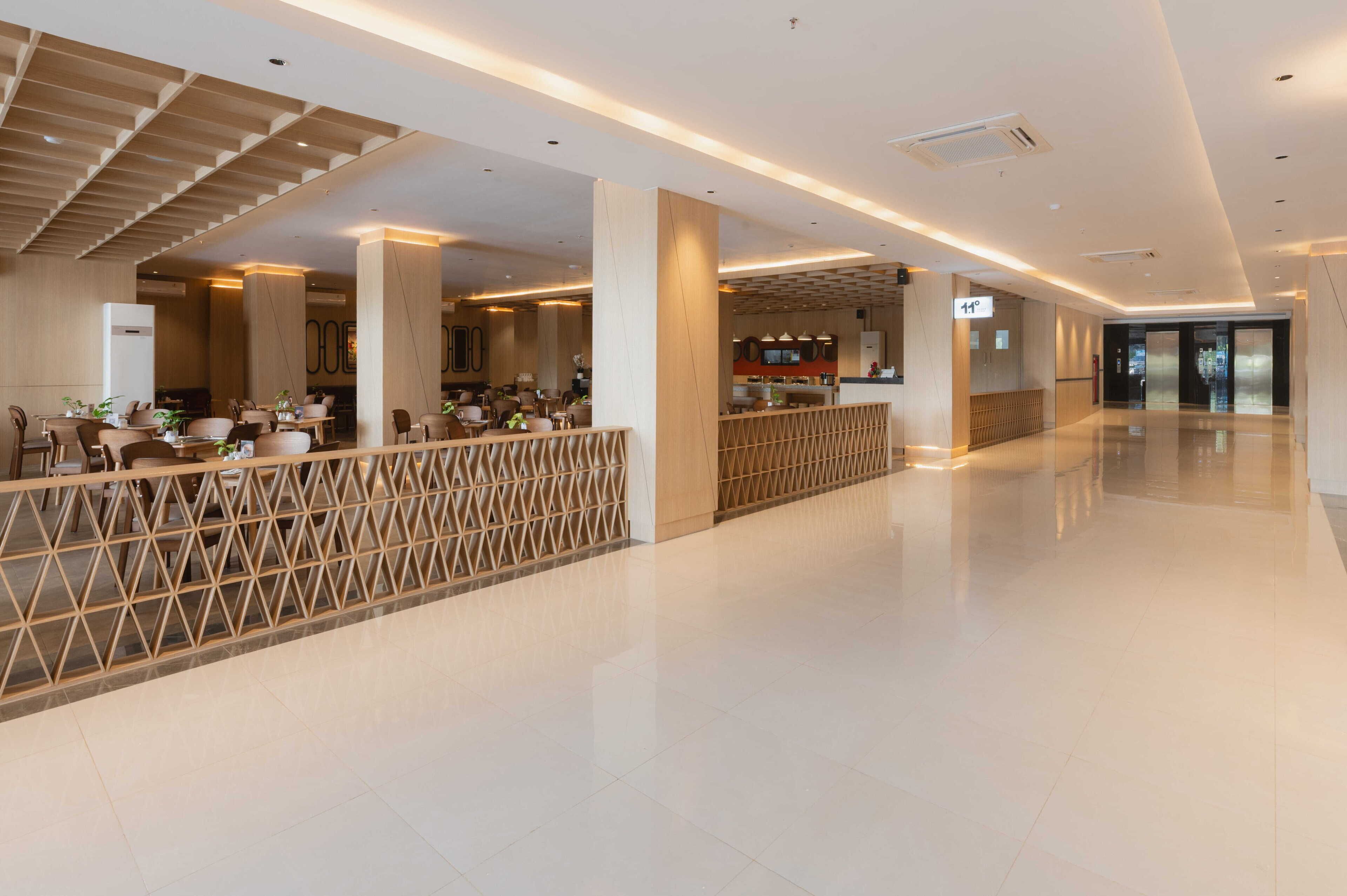 A spacious hotel lobby with a modern design, featuring a central seating area with tables and chairs, a decorative wooden partition, and a clear view of the elevator area.