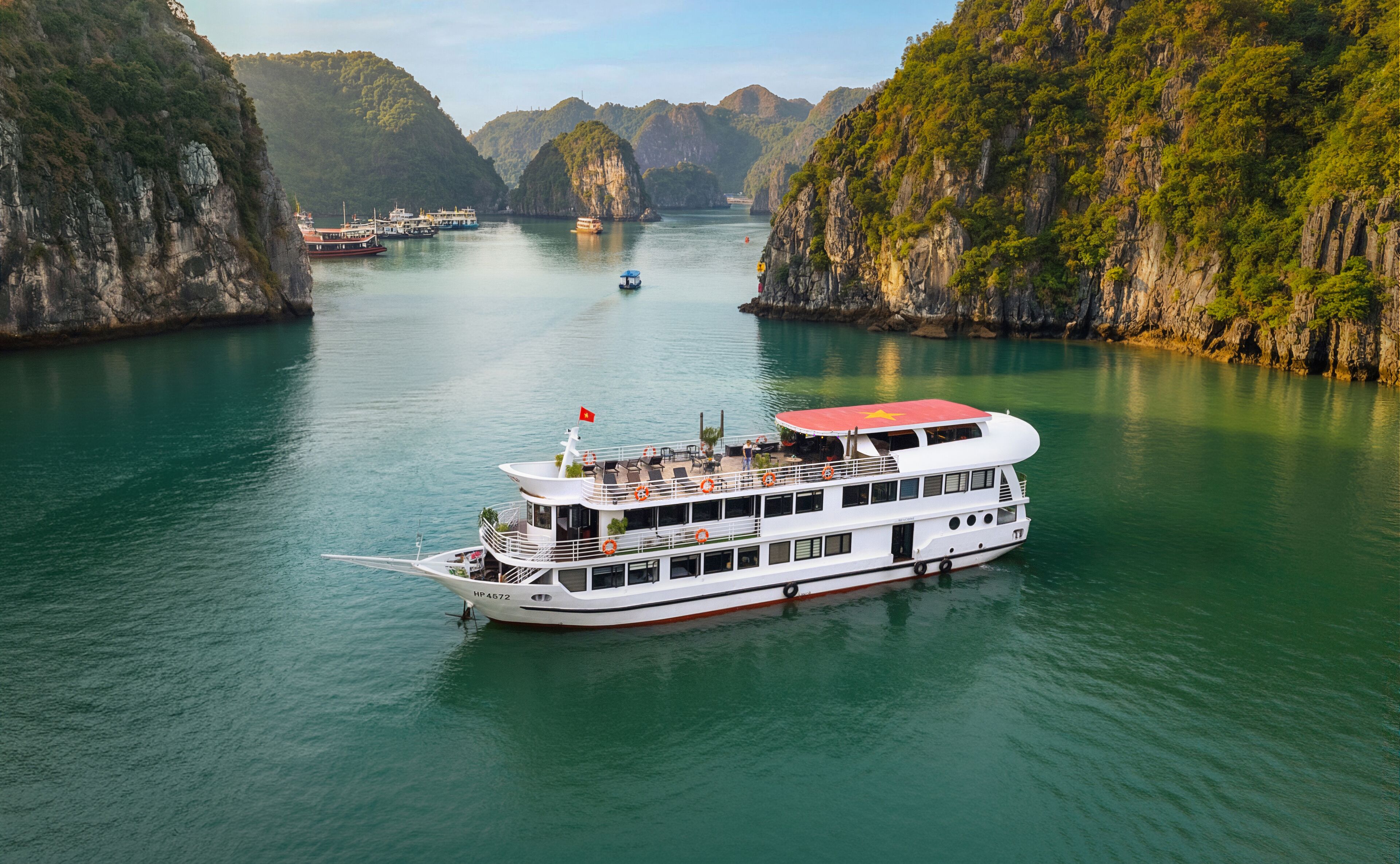 A large white riverboat with multiple decks and a red roof, docked in a calm river surrounded by steep, forested cliffs.