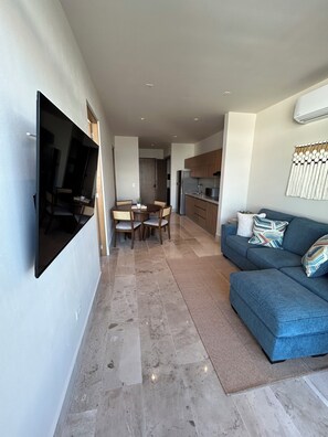 A modern living space with a flat-screen TV mounted on the wall, a dining area with a table and chairs, a blue sofa with patterned cushions, and a kitchen area with wooden cabinets and a refrigerator. - Brand New Condo In Beautiful Cozumel - 2 Bedroom/2 Bath With AC and Fitness Room (San Miguel de Cozumel)