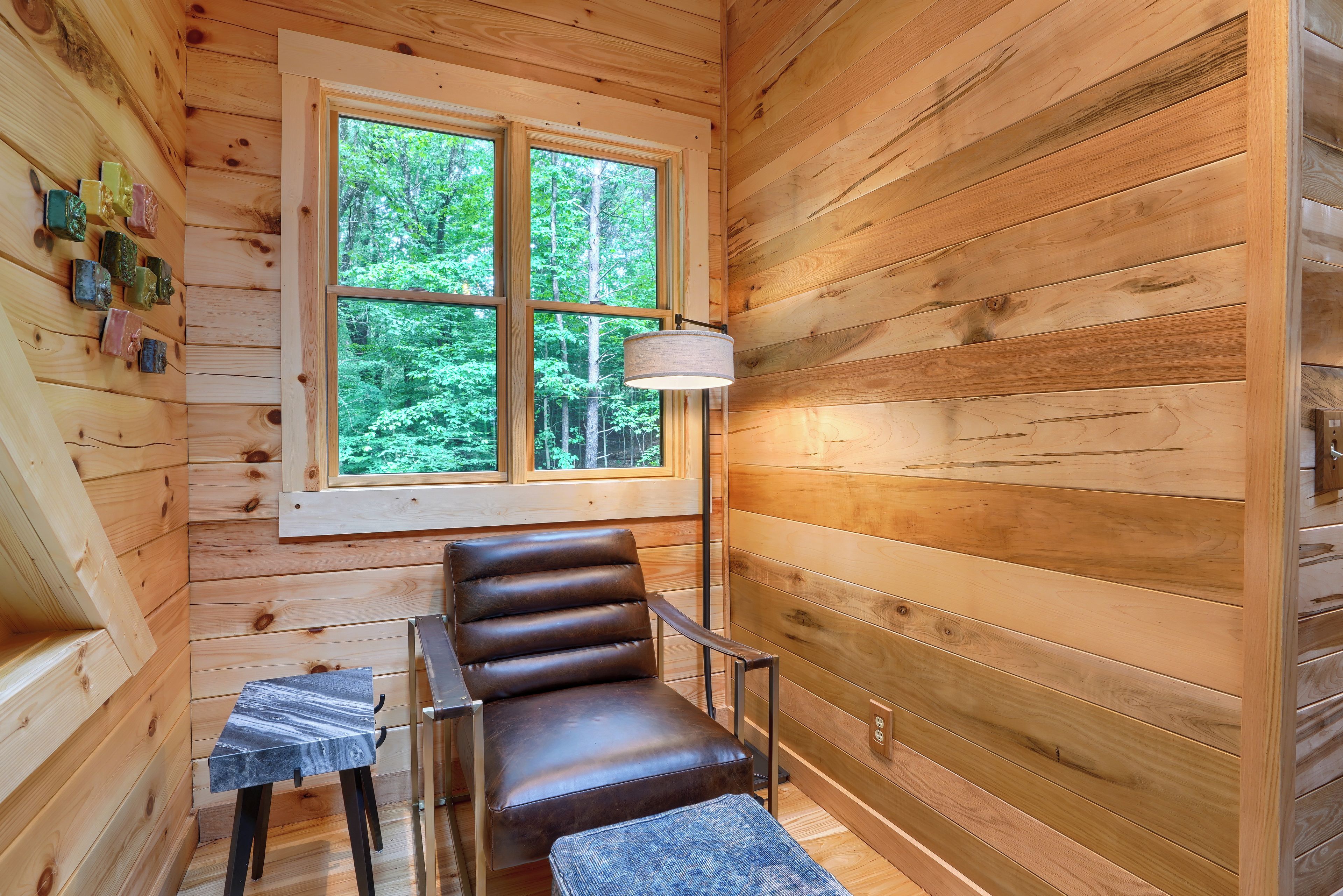 A wooden cabin interior with a leather chair, a small table, and a window with a view of greenery.
