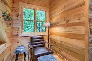 A wooden cabin interior with a leather chair, a small table, and a window with a view of greenery. - Beehive Bungalow *Charming Forest Escape* Hot Tub (Logan)