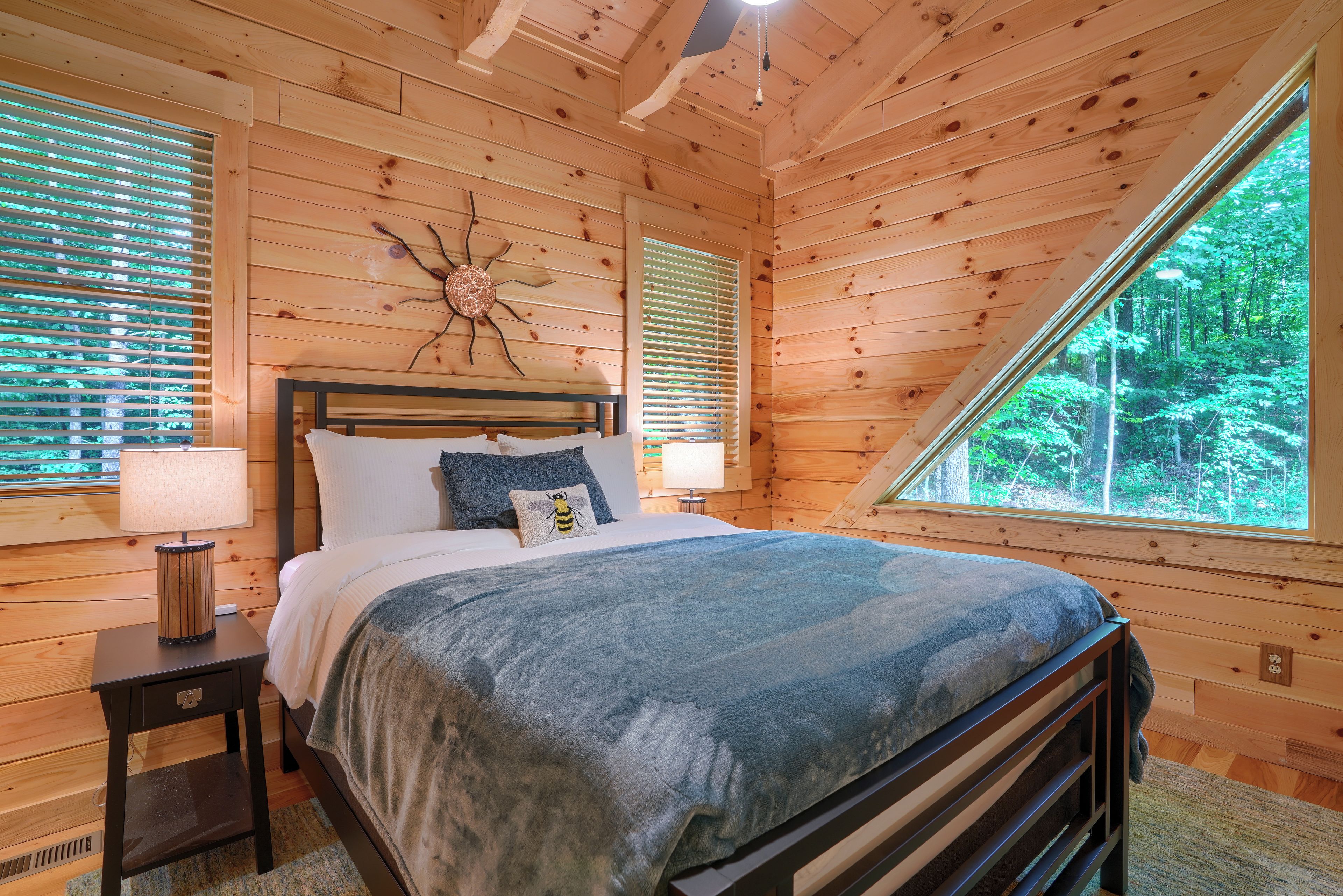 A bedroom with a wooden ceiling, a bed with a blue blanket, two bedside lamps, and a window with blinds.