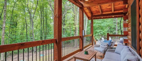 Terraza de madera con un sofá y una mesa, rodeada por barandilla de madera, con vista a un bosque.