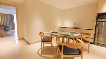 A modern kitchen with a stainless steel refrigerator, a dining table with chairs, and a view into a living area with a sofa and curtains.