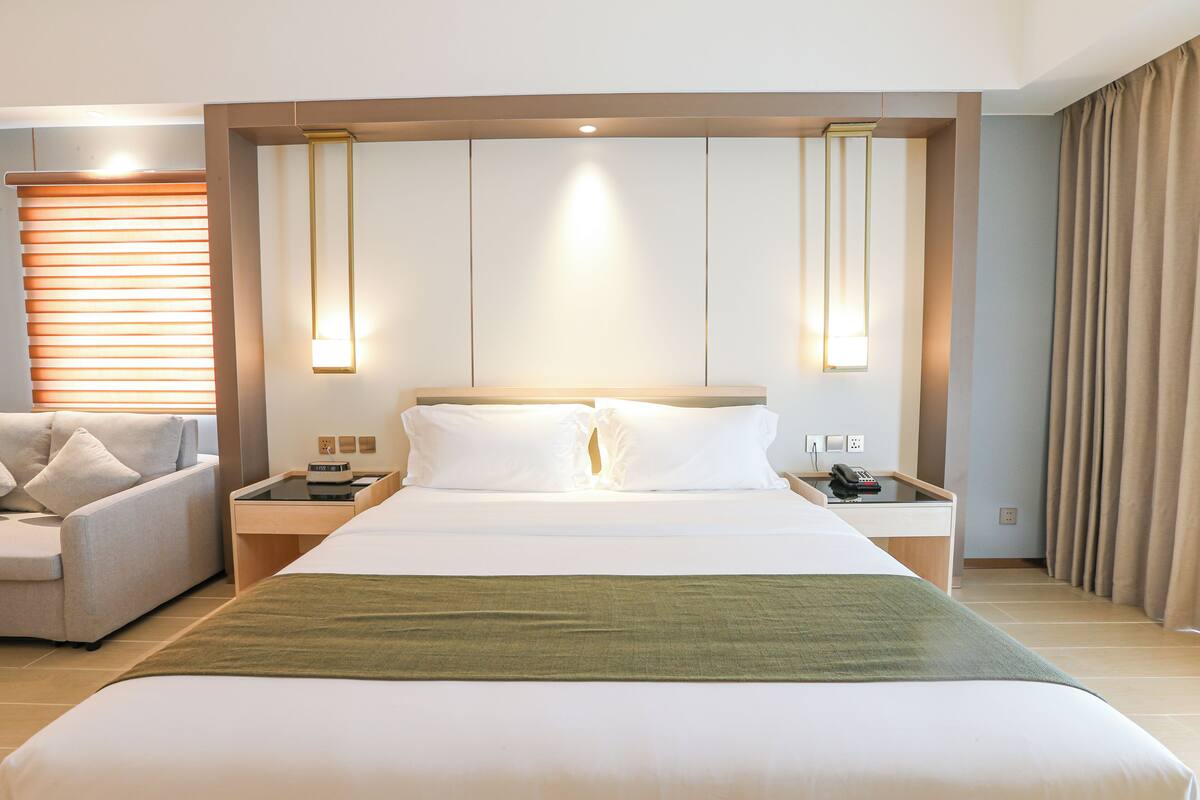 Ein Hotelzimmer mit einem großen Bett, einem Nachttisch mit Telefon und Lampe, einem Sofa und einem Fenster mit Jalousien.