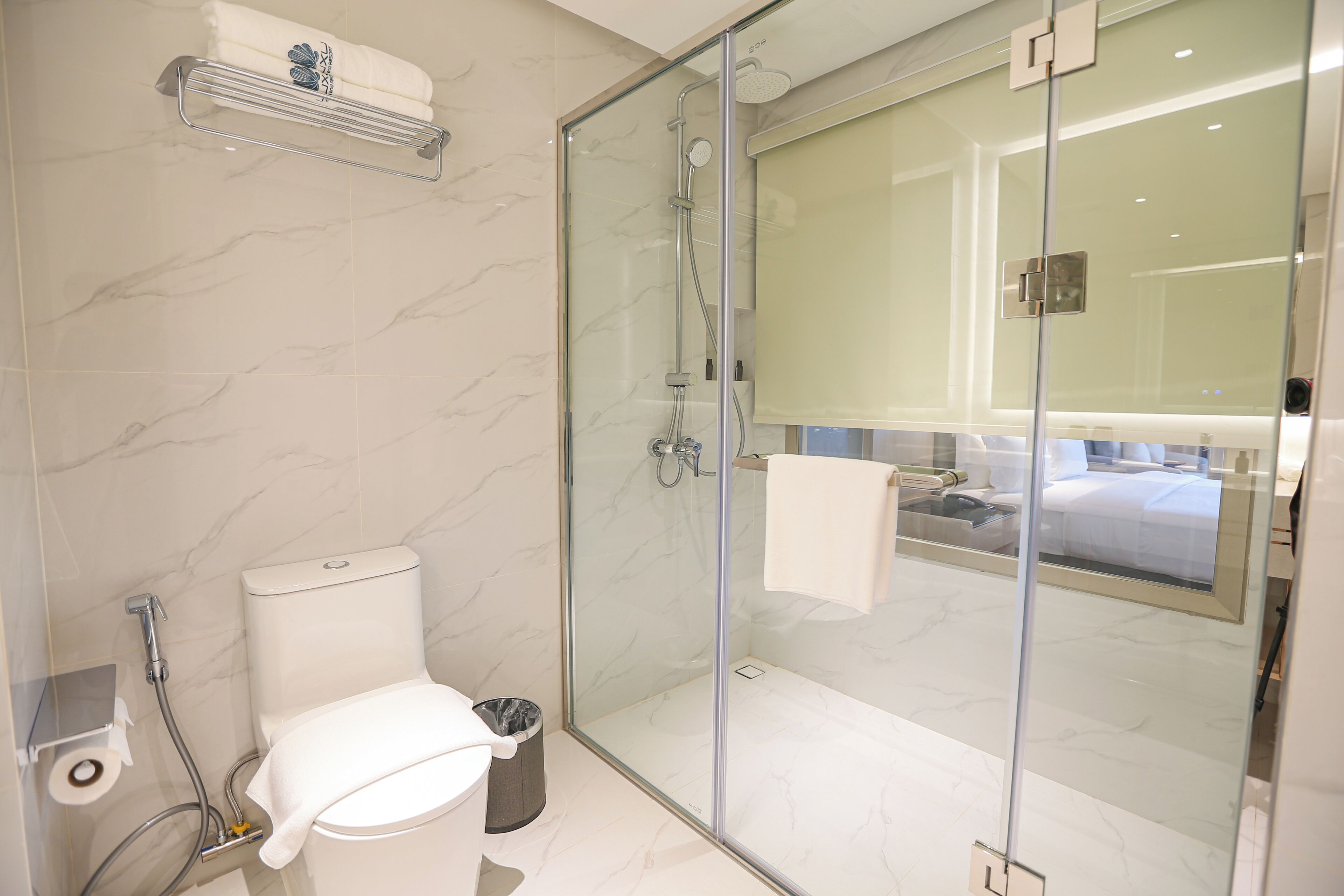 A modern bathroom with a glass shower enclosure, a toilet, and a visible bed in the adjacent room.