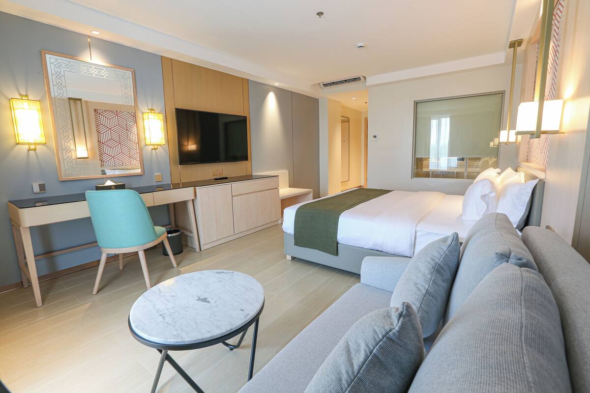 Ein modernes Hotelzimmer mit Bett, Sofa, Schreibtisch, Sessel, Fernseher und einem kleinen Tisch.