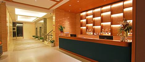 A hotel reception area with a wooden reception desk, potted plants, and a staircase leading to an elevator.